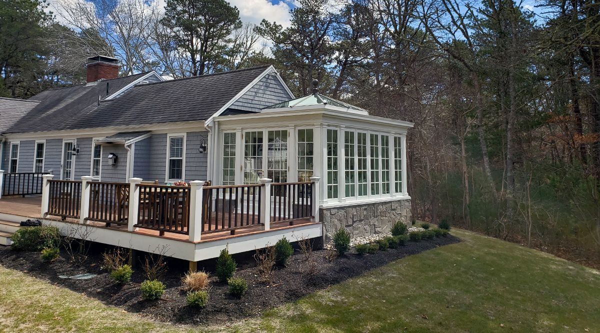 Light blue house with a sunroom, deck, and stone base, surrounded by trees and a lawn.