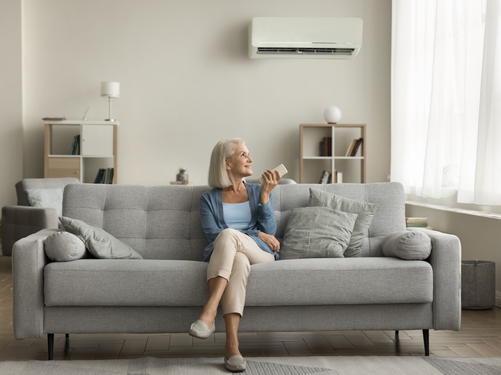 Woman sits on a gray sofa in a living room, using an air conditioner remote.