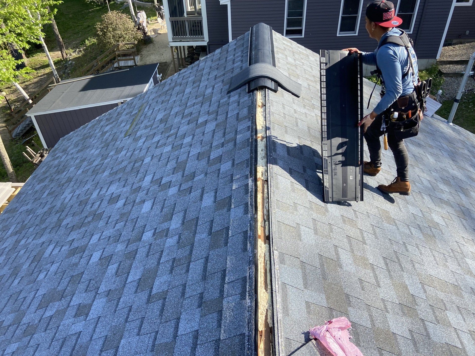 Person on rooftop installing vent. Roof is gray with some trees in background.