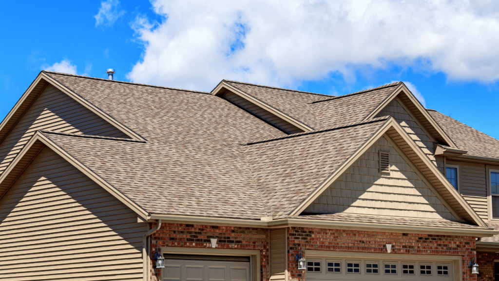 Tan shingled roof of a house against a blue sky with white clouds.