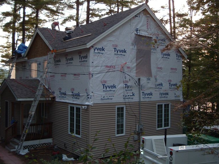 House under construction, with roofers working on the roof and workers on a ladder. House covered in Tyvek.