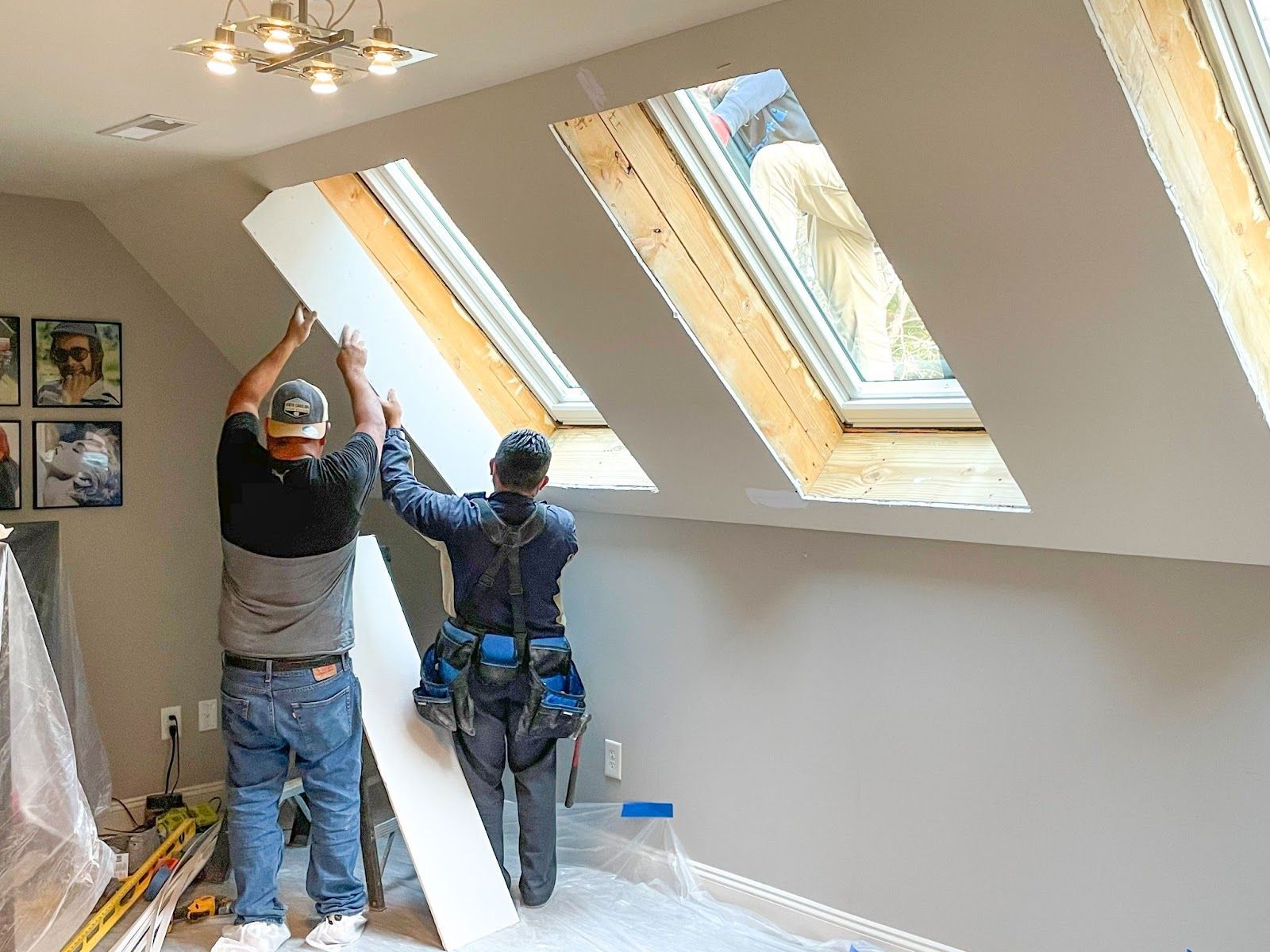 Two people installing white drywall near angled windows in a room.