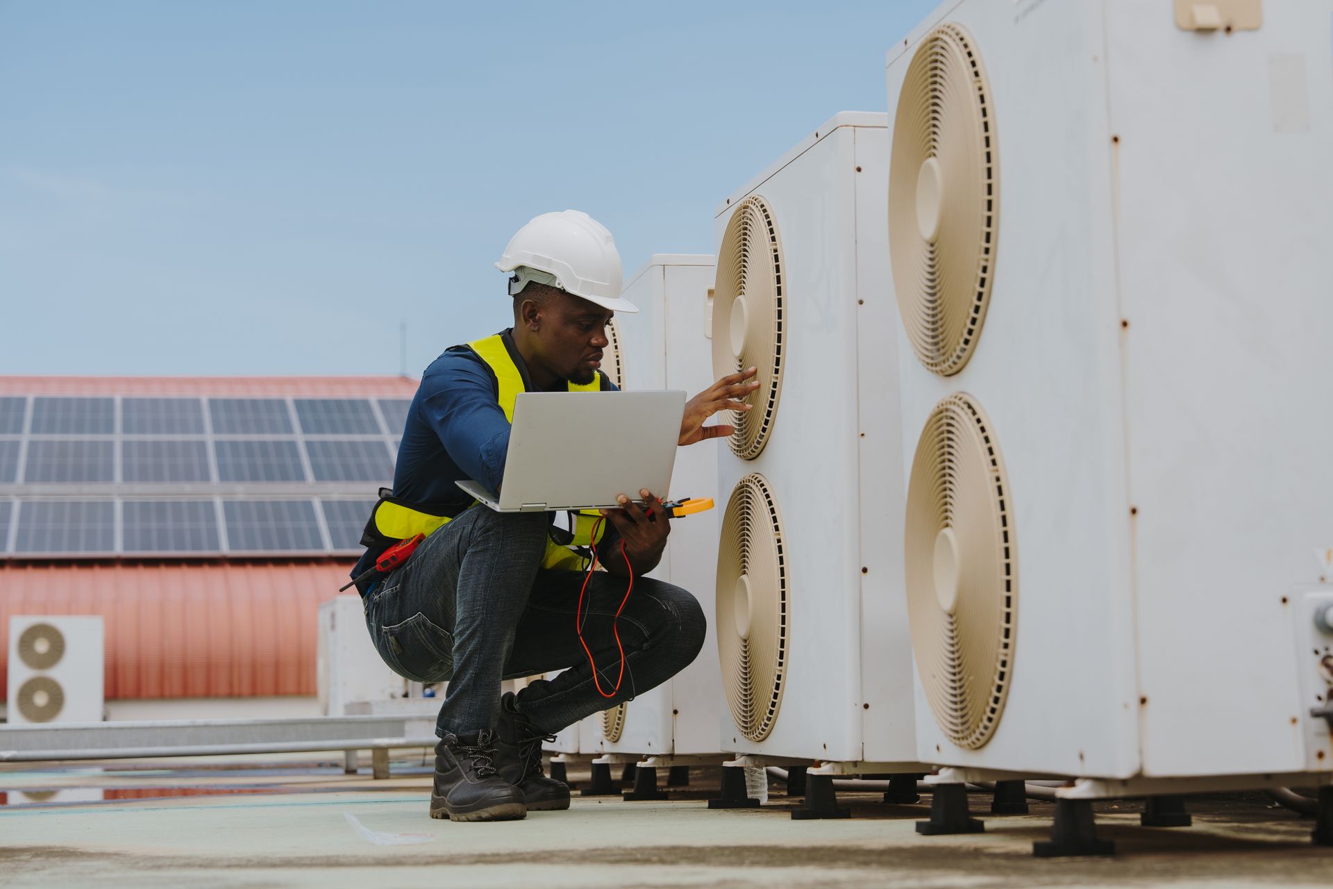 HVAC technician in hard hat inspects rooftop air conditioning units with laptop and multimeter.