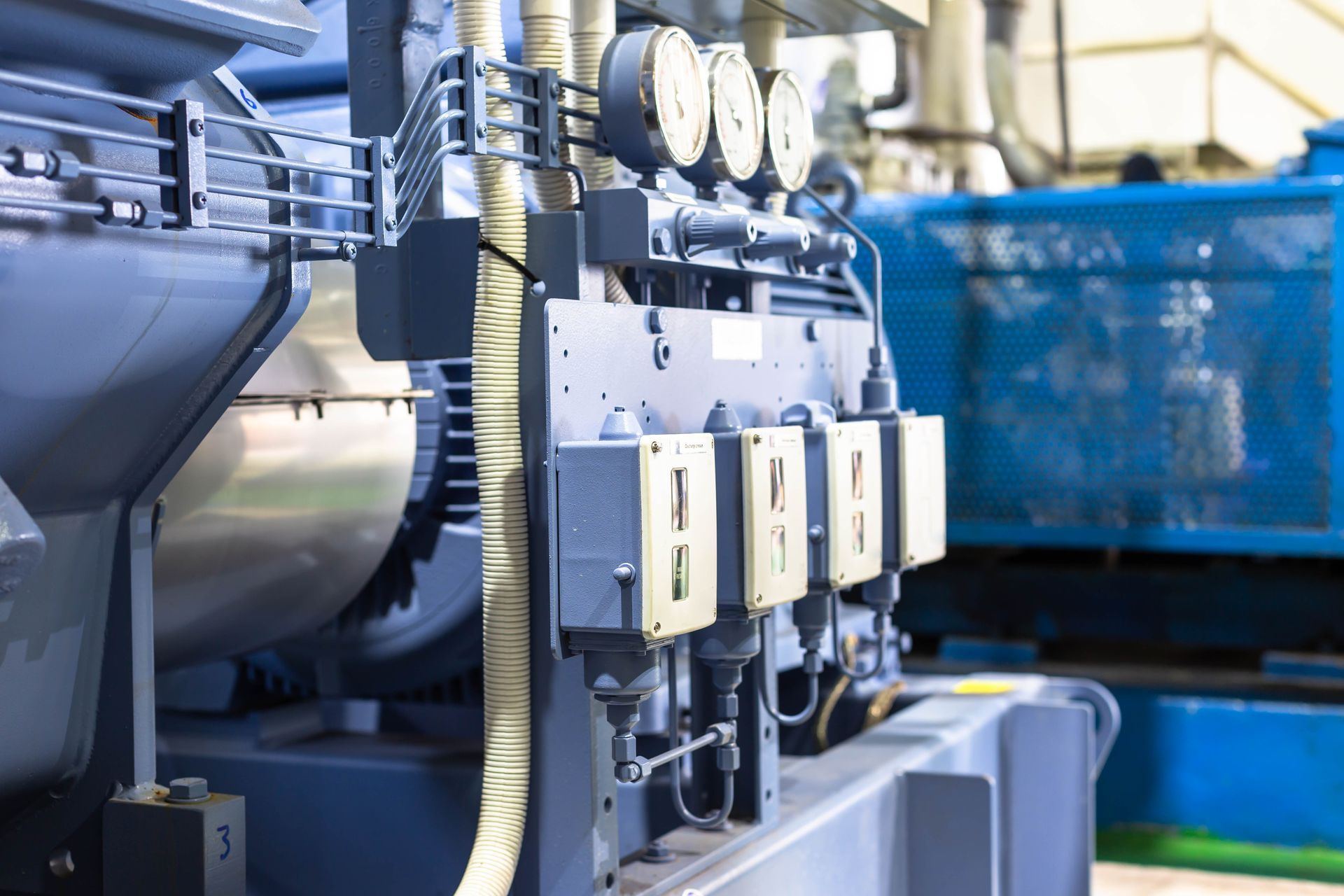 Close-up of industrial machinery: gauges, electrical boxes, and pipes on a large, blue-gray machine in a factory setting. Close-up of industrial machinery: gauges, electrical boxes, and pipes on a large, blue-gray machine in a factory setting.