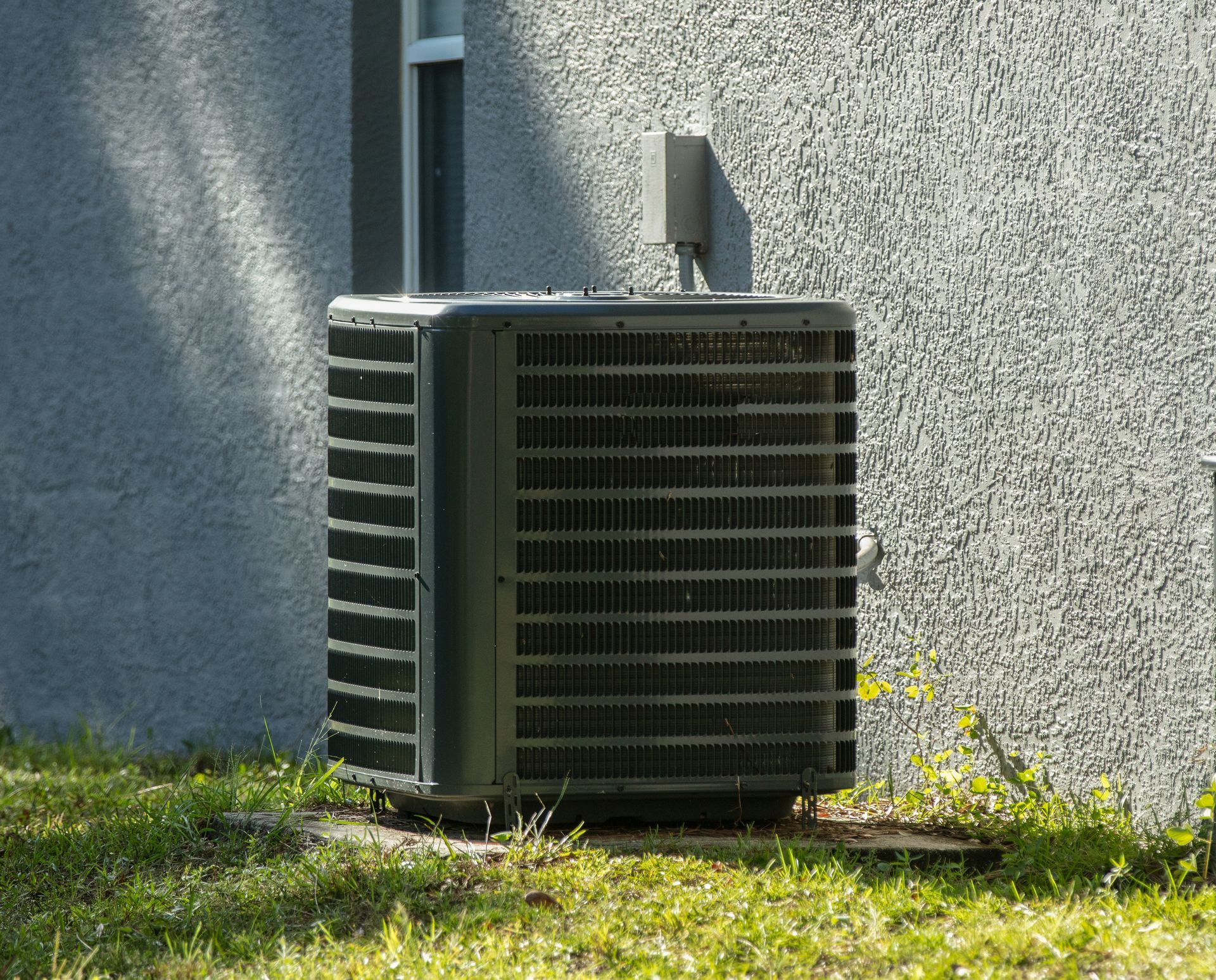 Hands repairing an air conditioning unit with tools; white unit, red and black plaid shirt.