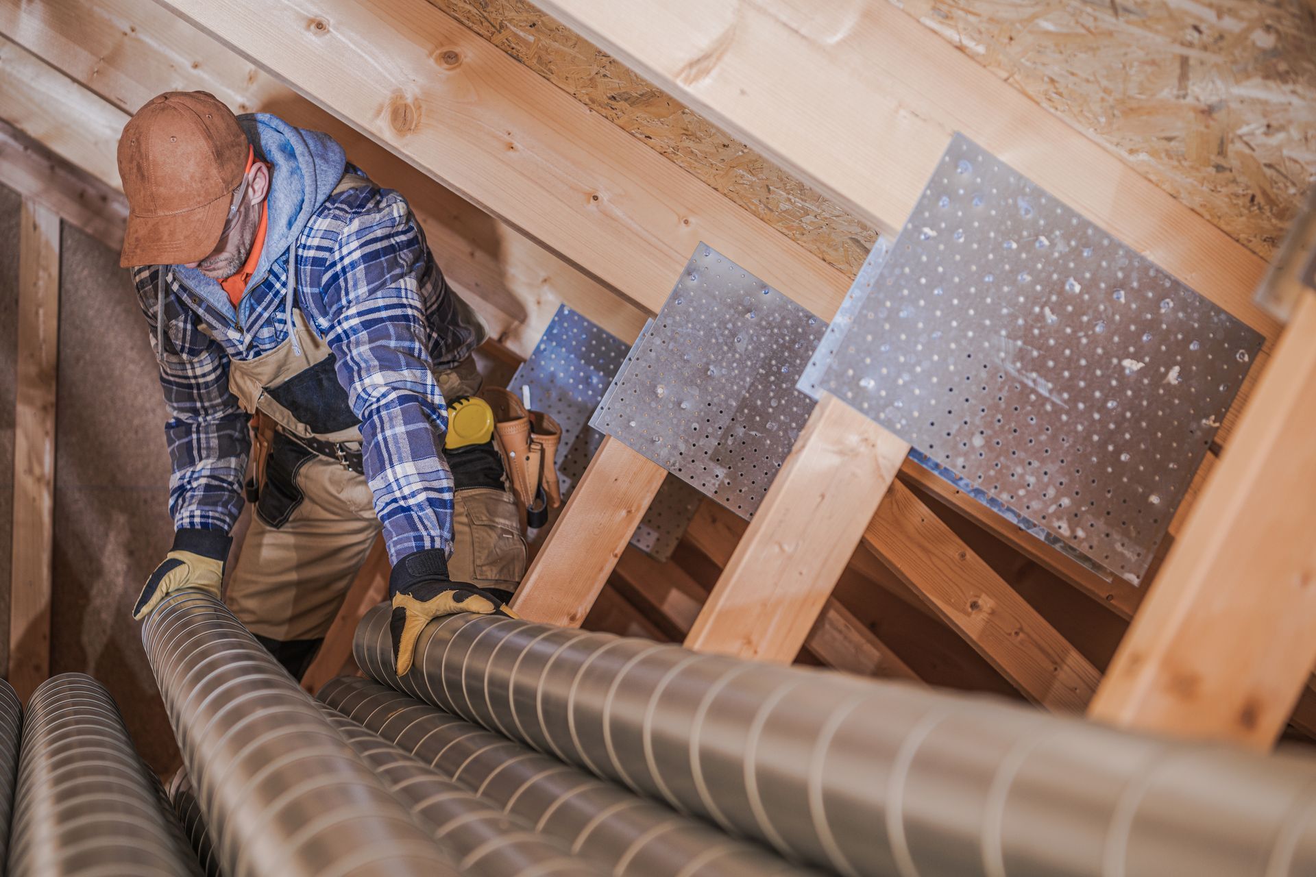 Construction worker in an attic installing HVAC ductwork, wooden beams, metallic tubing, wearing gloves and a hard hat. Construction worker in an attic installing HVAC ductwork, wooden beams, metallic tubing, wearing gloves and a hard hat.