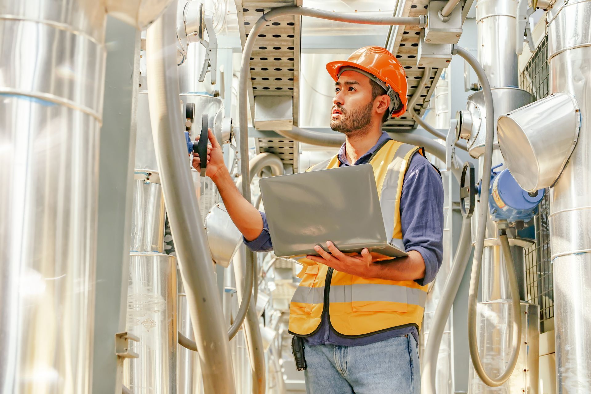 Engineer in hard hat and safety vest, using a laptop in an industrial facility, surrounded by pipes. Engineer in hard hat and safety vest, using a laptop in an industrial facility, surrounded by pipes.