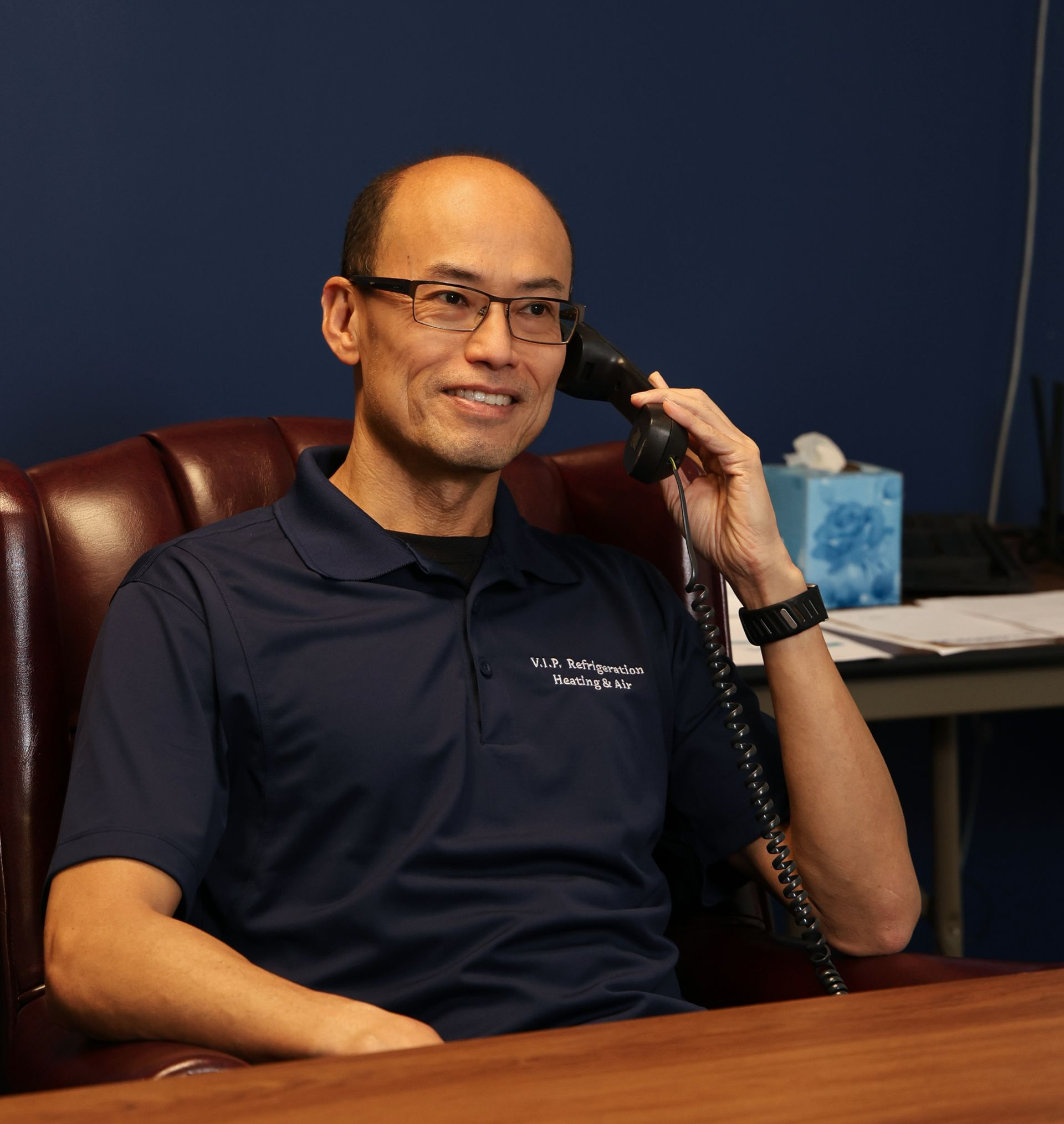 Man in glasses on phone, smiling in office. Dark blue shirt, brown leather chair.