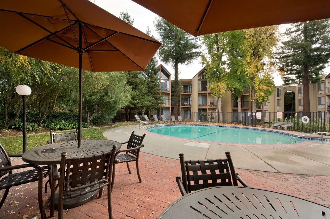 Poolside seating area with umbrellas, tables, and chairs overlooking a pool and apartment buildings.