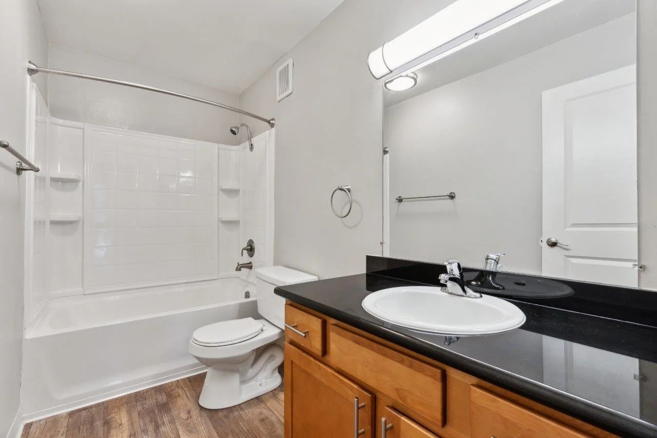 Bathroom with a white tub/shower combo, toilet, sink, and dark countertop; wooden cabinets.