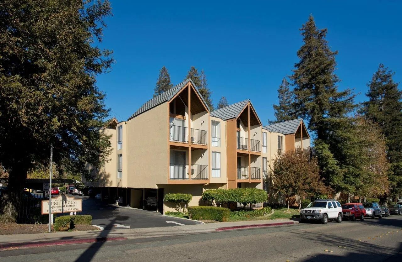 Apartment building with beige walls and dark wood trim, trees in the background, clear blue sky.