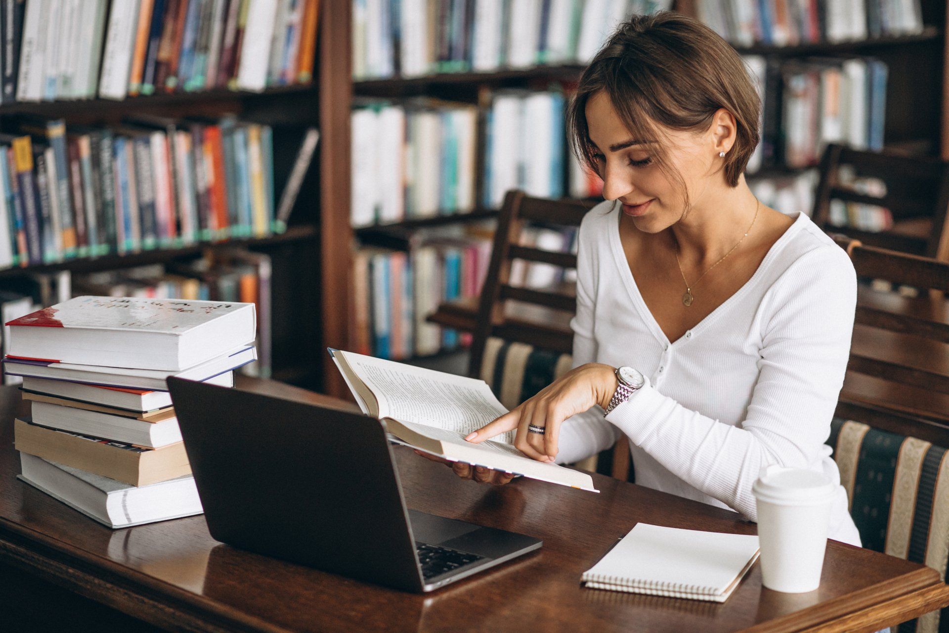 een vrouw zit aan een tafel in een bibliotheek, leest een boek en gebruikt een laptop.