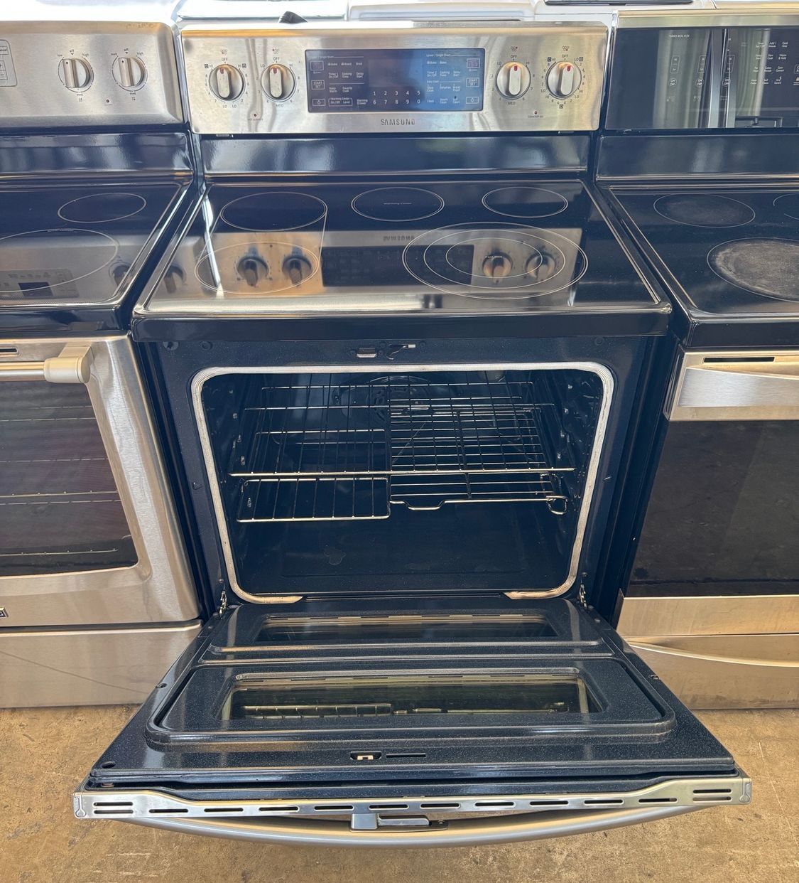 A stainless steel oven with the door open, showcasing the interior racks. It's in a row of similar appliances.