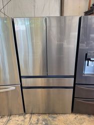 A stainless steel four-door refrigerator stands among other appliances. It has a top section with two doors and two freezer drawers below.