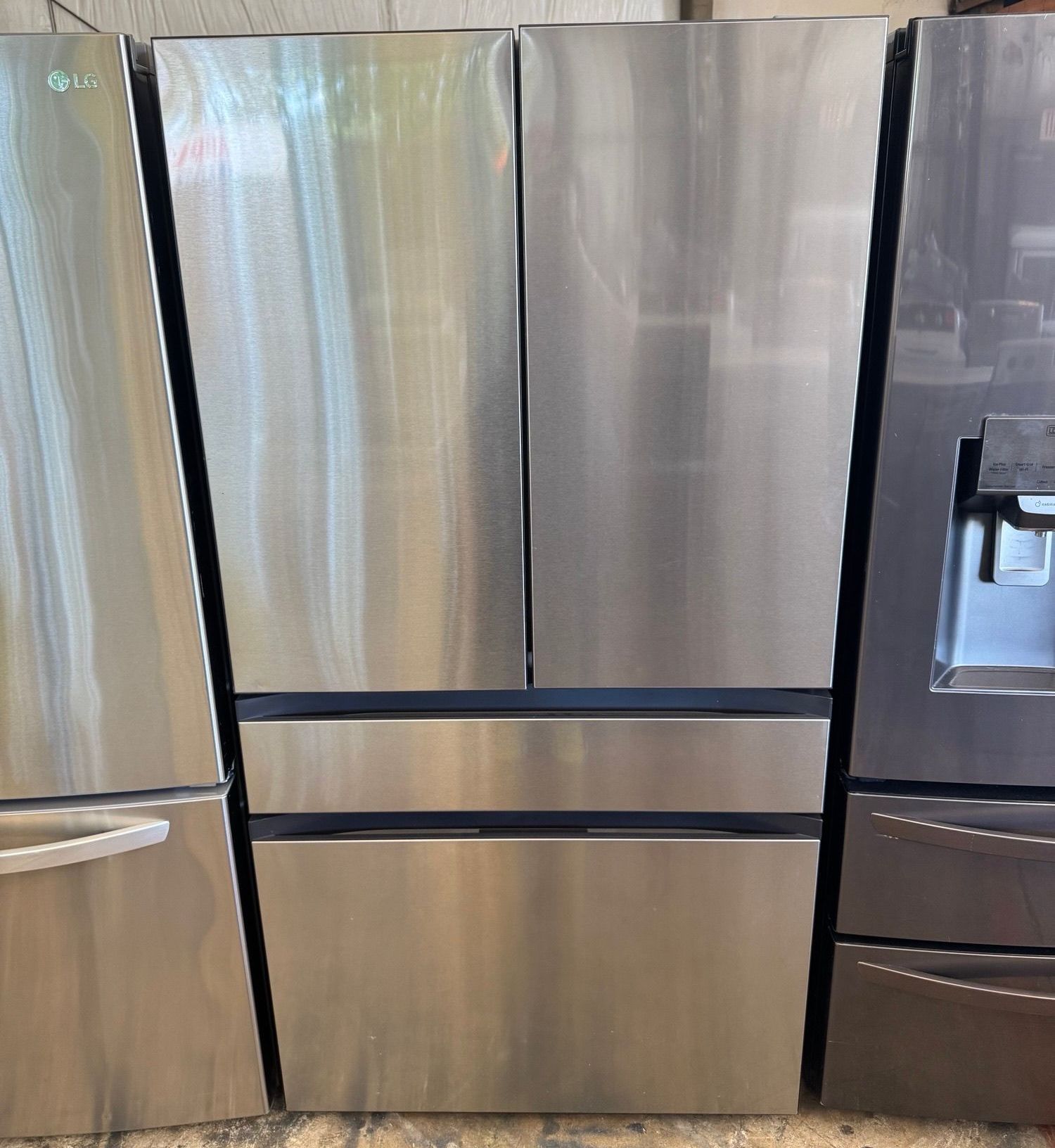 Four-door stainless steel refrigerator with two upper doors and two lower drawers, standing among other appliances.