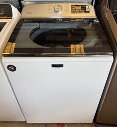 A white top-load washing machine with a glass lid and silver control panel. It is located between other appliances.