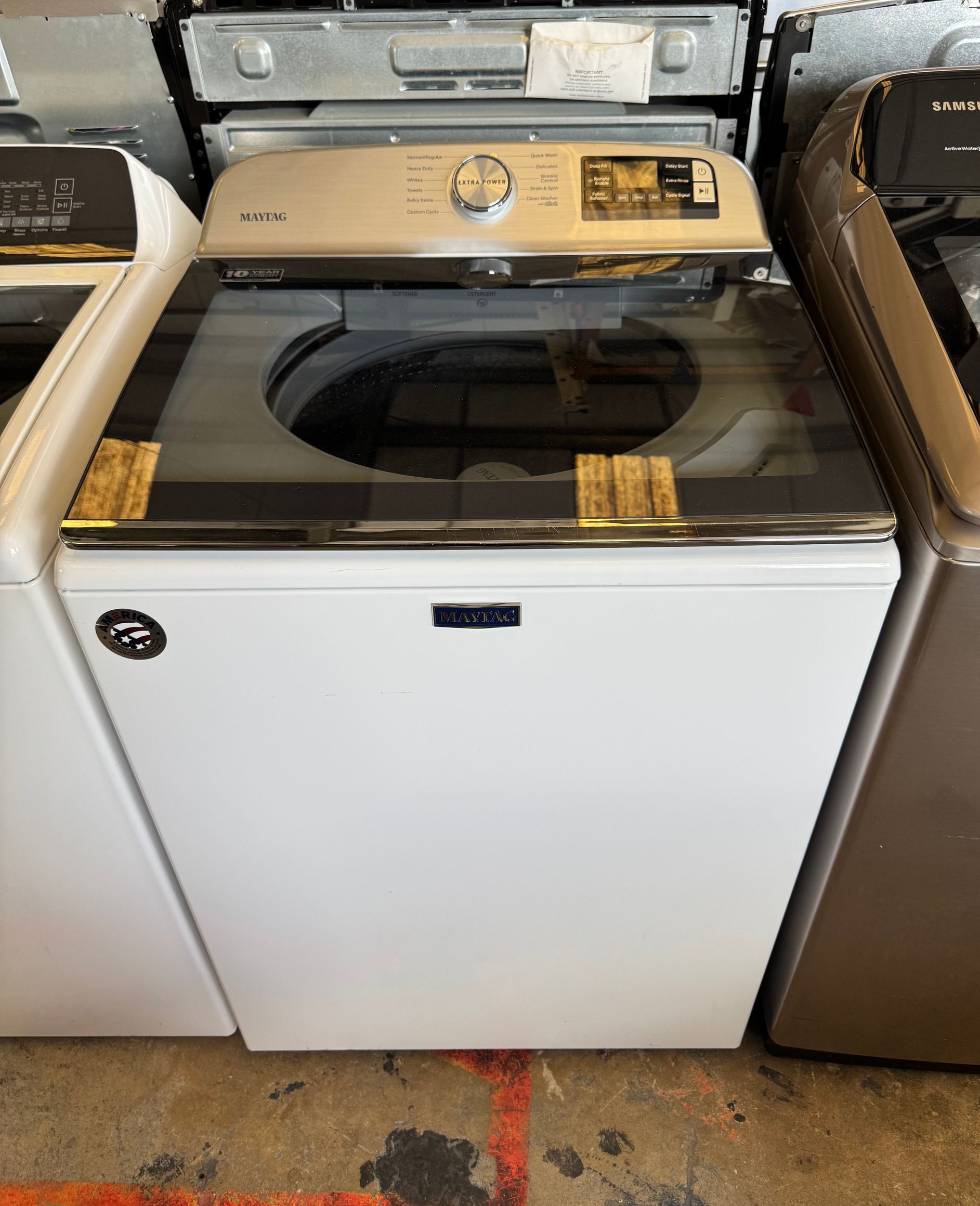 A white top-load washing machine with a glass lid and silver control panel. It is located between other appliances.