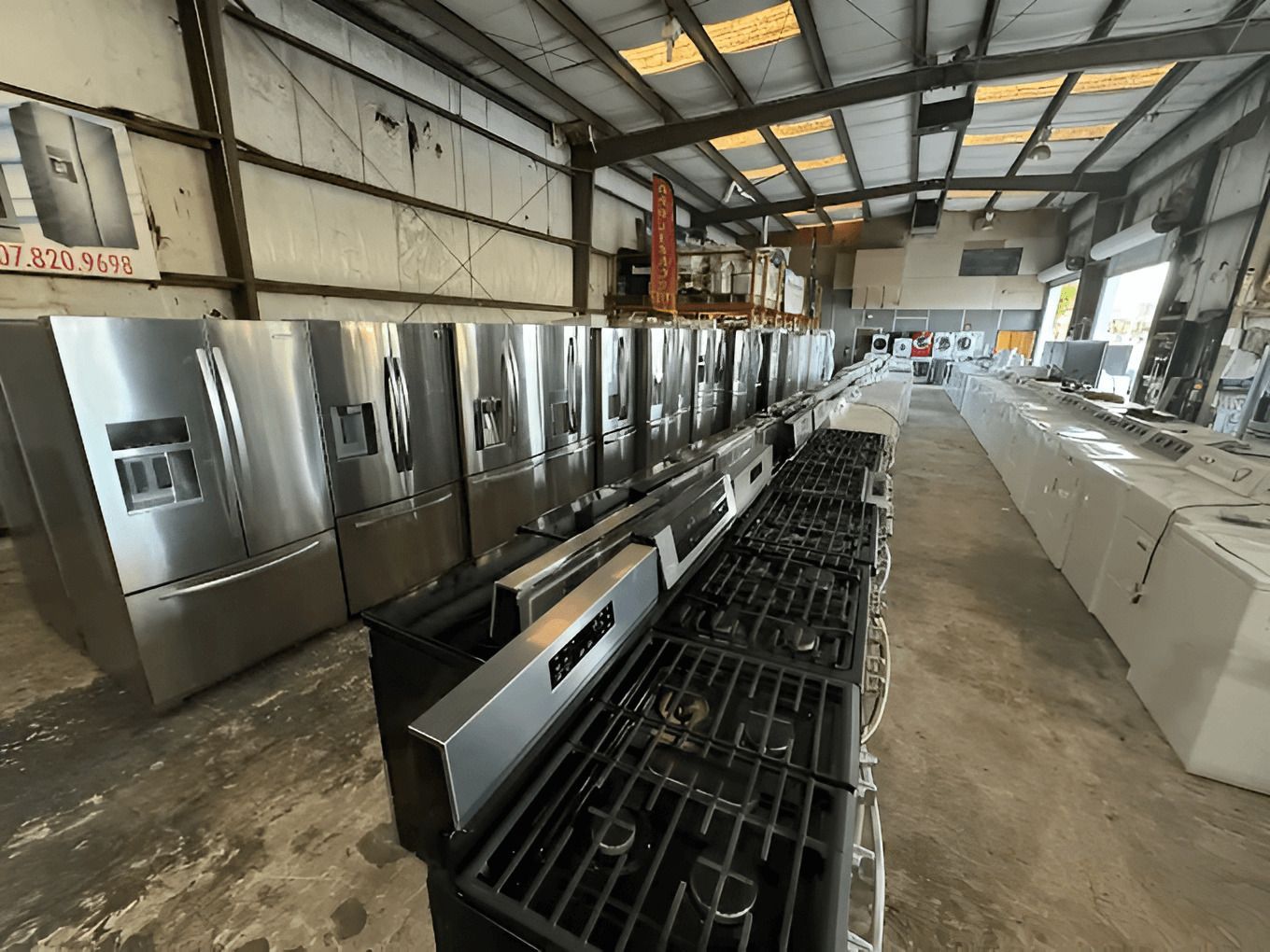 A warehouse displays rows of stainless steel refrigerators and gas ranges, with white countertops and other appliances further in the background.