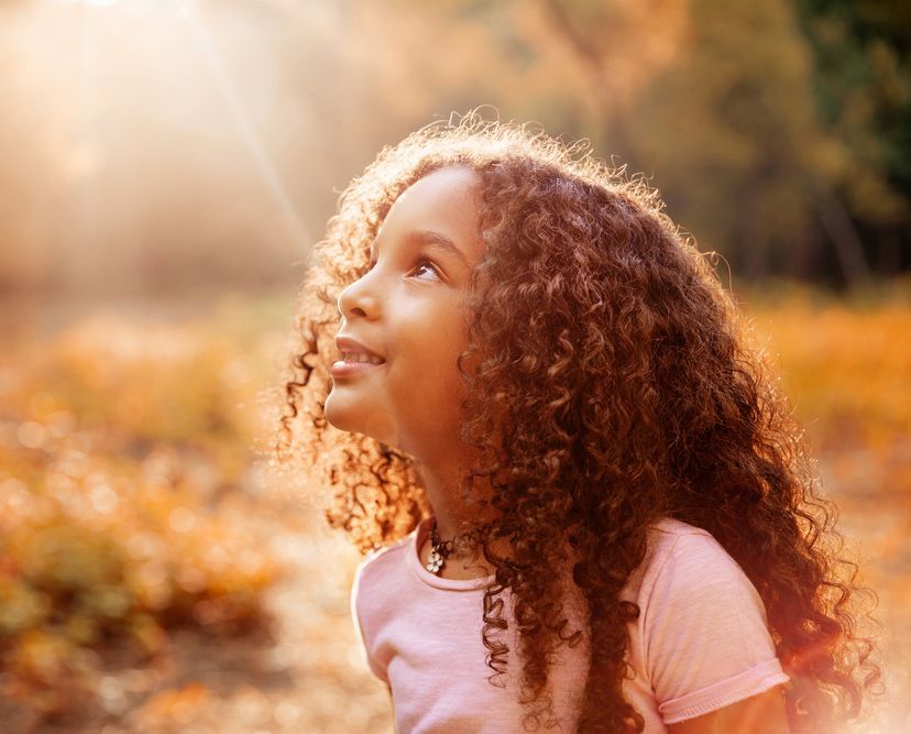 A little girl with curly hair is looking up at the sun