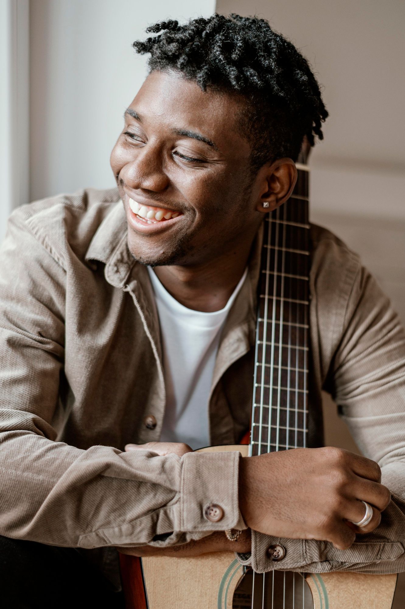 Smiling Black man with guitar, leaning against a window, wearing a tan shirt and a white shirt.