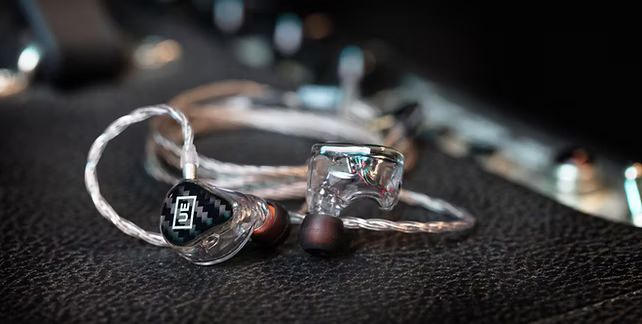 Clear in-ear monitors with silver cable resting on a textured dark surface. One earbud has a black faceplate.