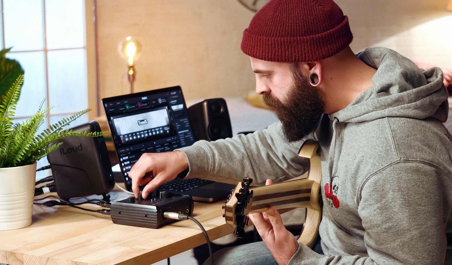 Man with beard and beanie plays guitar, recording on a laptop.