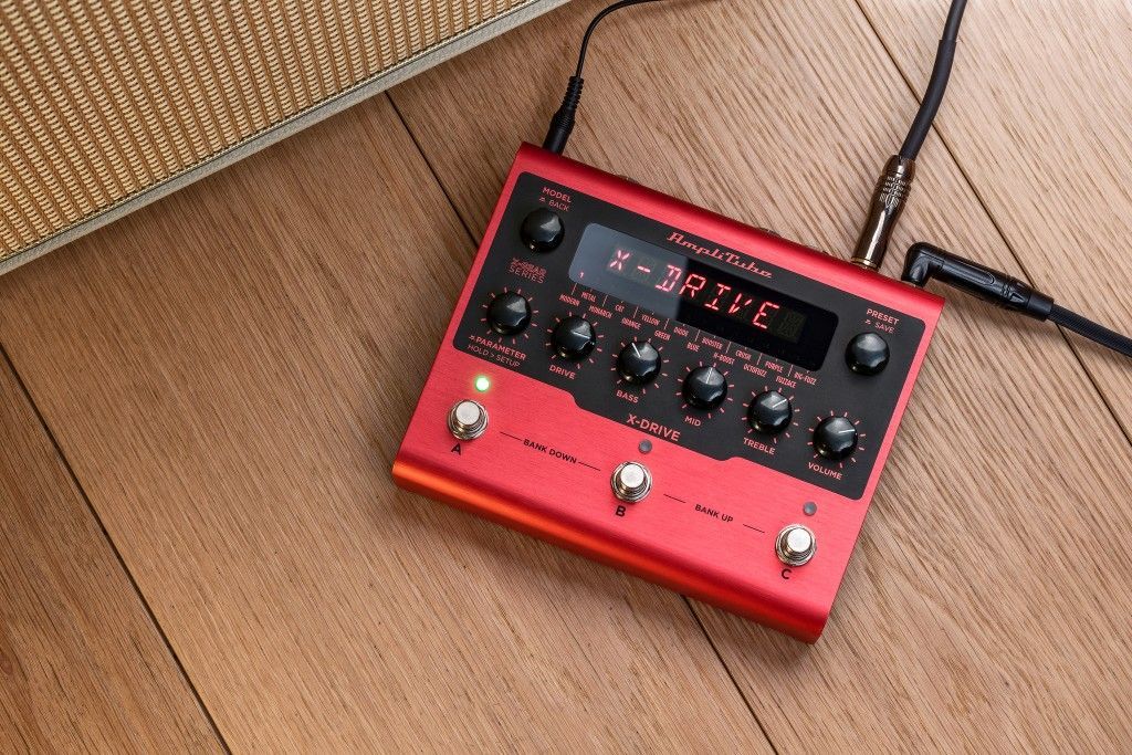 Red guitar effects pedal on wooden floor, with knobs, display, and connected cables.