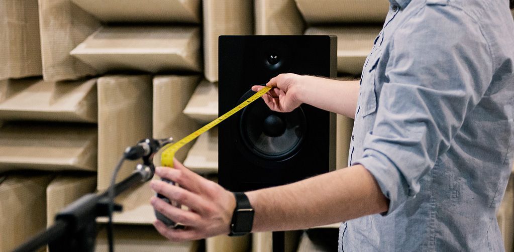 Person measuring a black speaker with a yellow tape measure in a soundproof room.