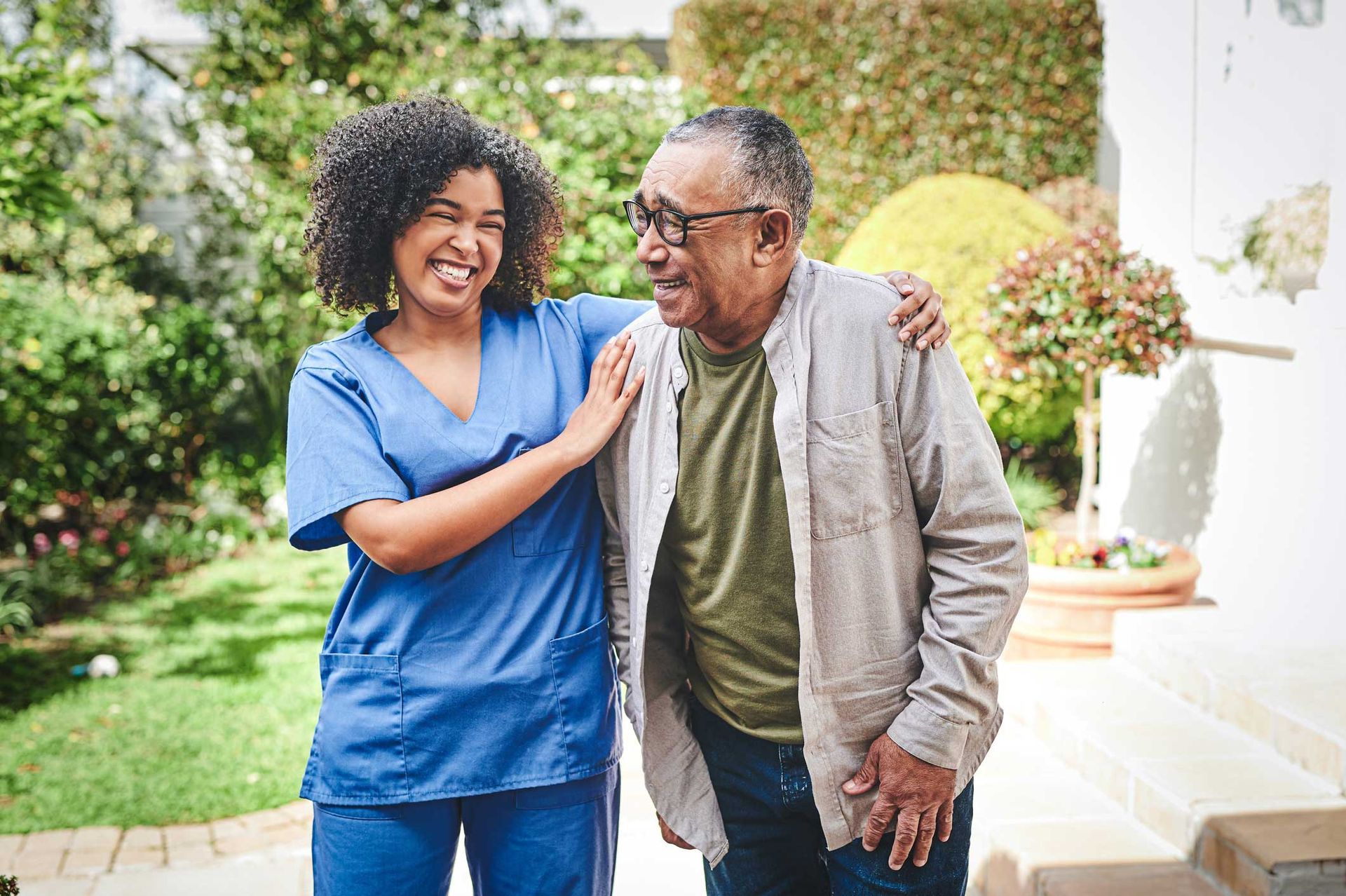 Woman in scrubs helps an elderly man walk outdoors. They are smiling.