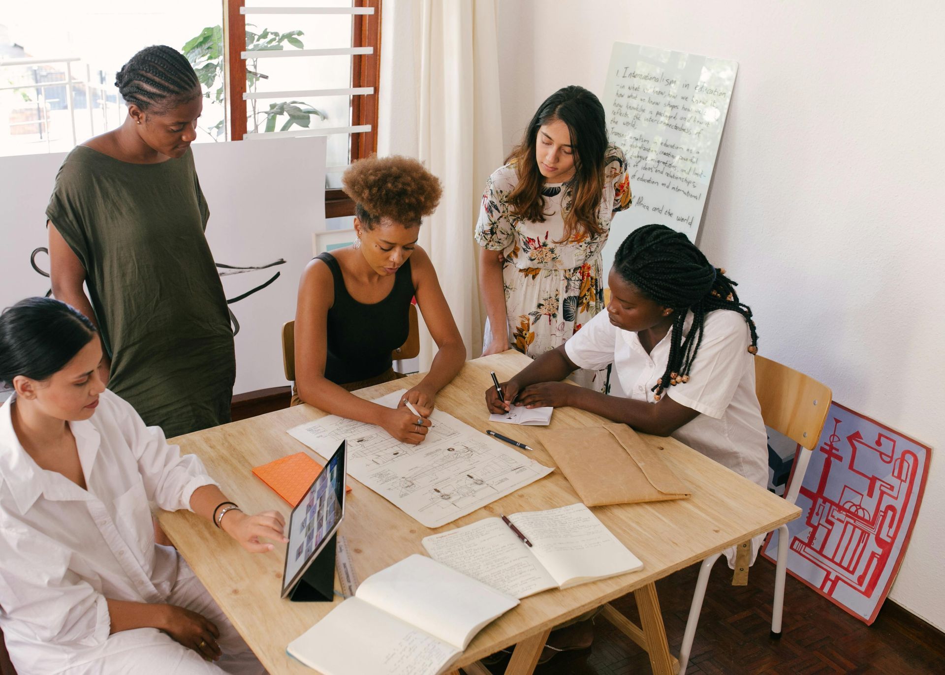 Group of women collaborating around a table with papers, a tablet, and notepads in a bright room.