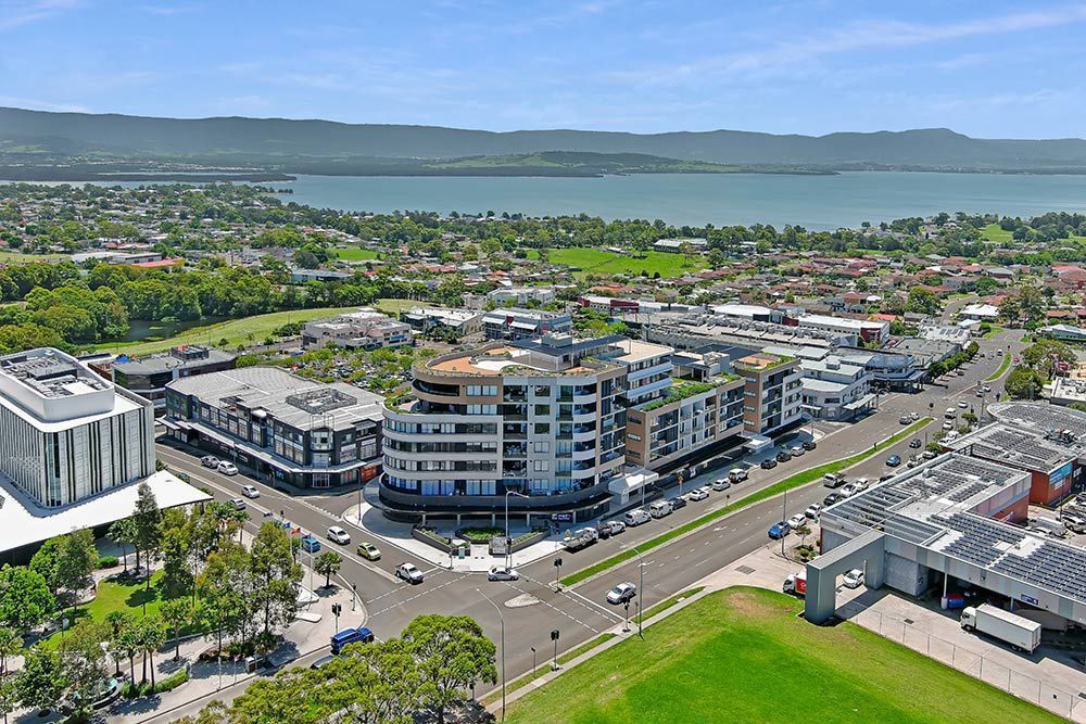 Aerial view of a town with modern buildings, green spaces, and a lake in the background on a sunny day.
