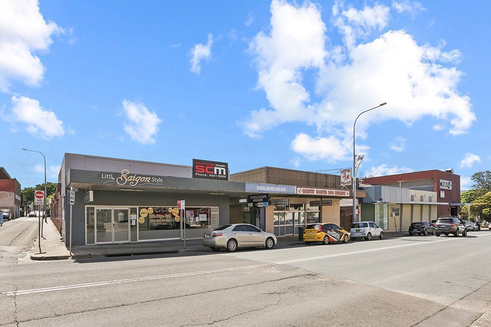 Street view of commercial buildings with shops and parked cars on a sunny day.