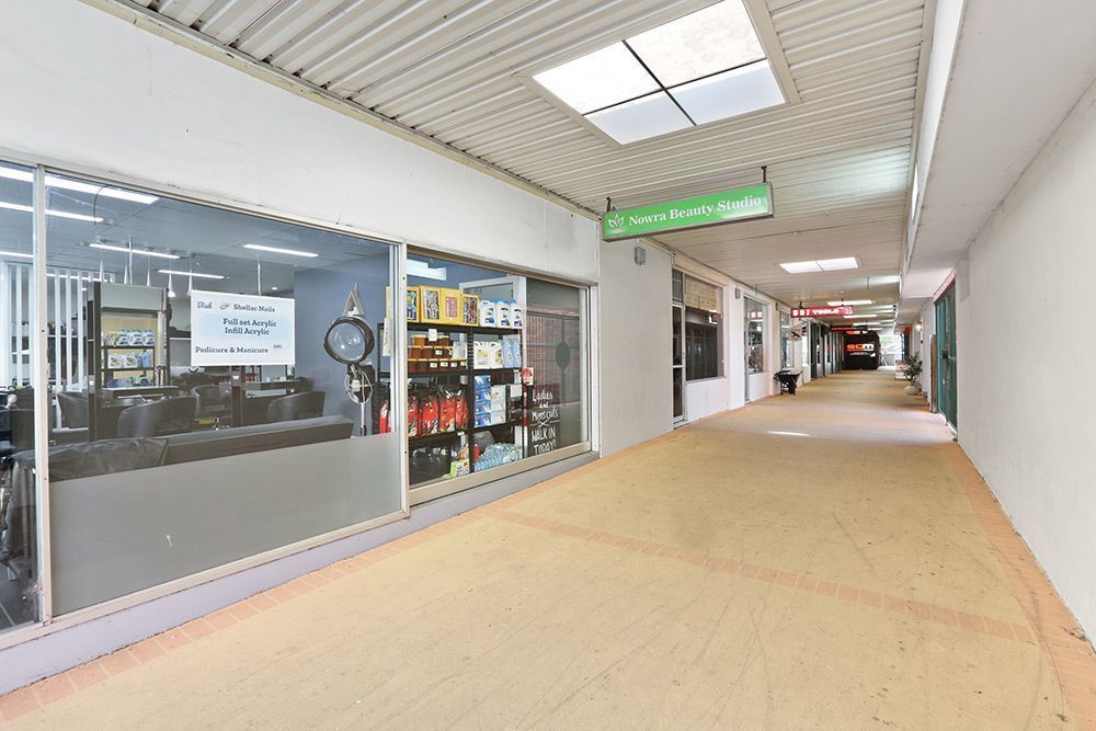 Long hallway in a commercial building with shops on the left, pale walls, and a bright ceiling with skylights.