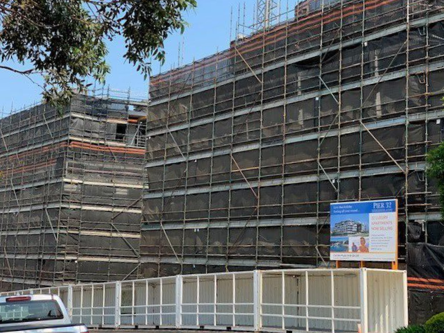Buildings under construction, covered in scaffolding and black netting; a sign advertising the project.