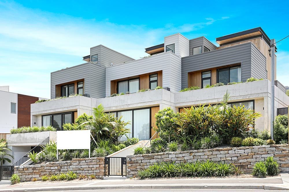 Modern multi-story home with neutral colors, lush landscaping, and blue sky.