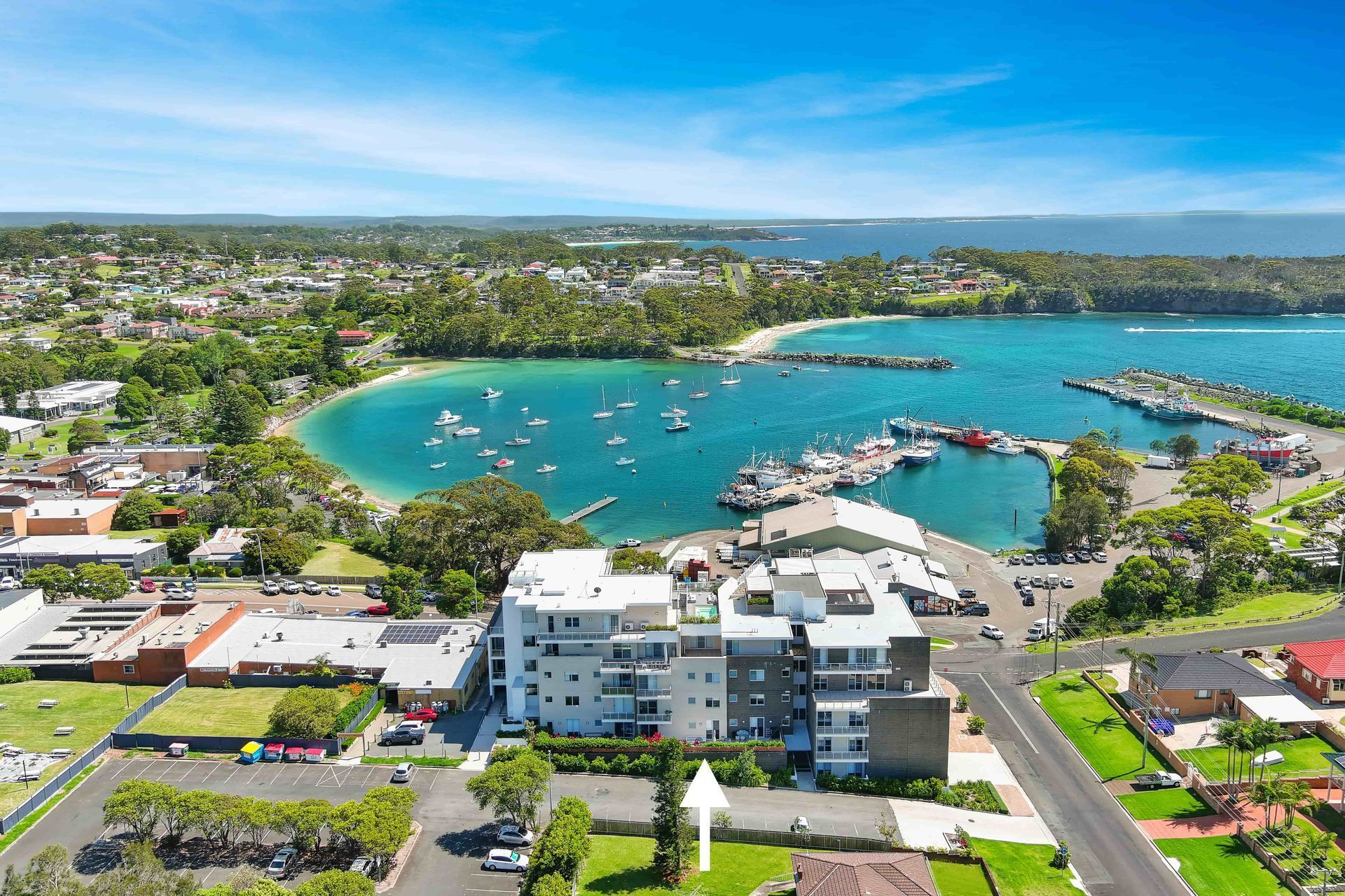 Aerial view: modern apartment building near harbor and town; boats on water, green trees.