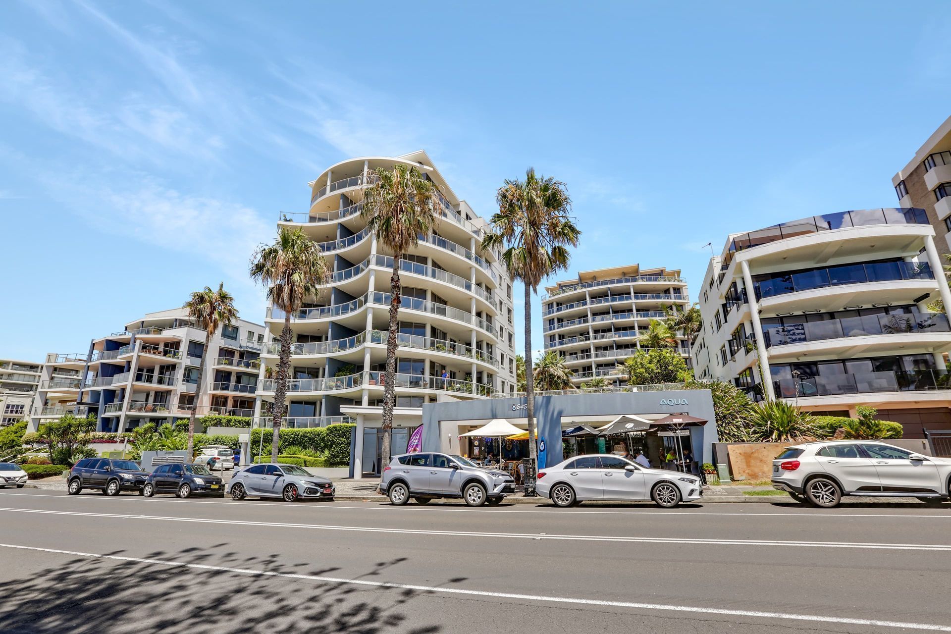 Apartment buildings with balconies line a sunny street, cars parked out front. Palm trees and blue sky.