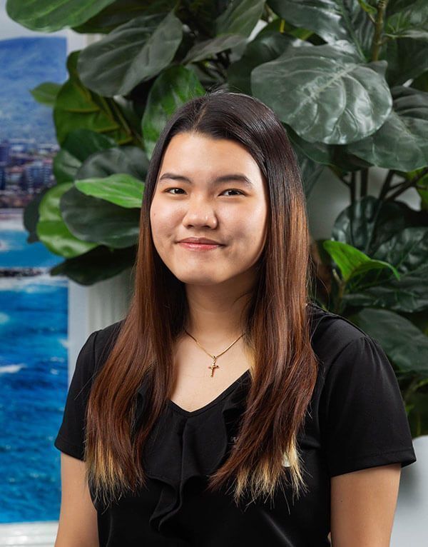 Young Asian woman with long brown hair, smiling in front of greenery and ocean backdrop.