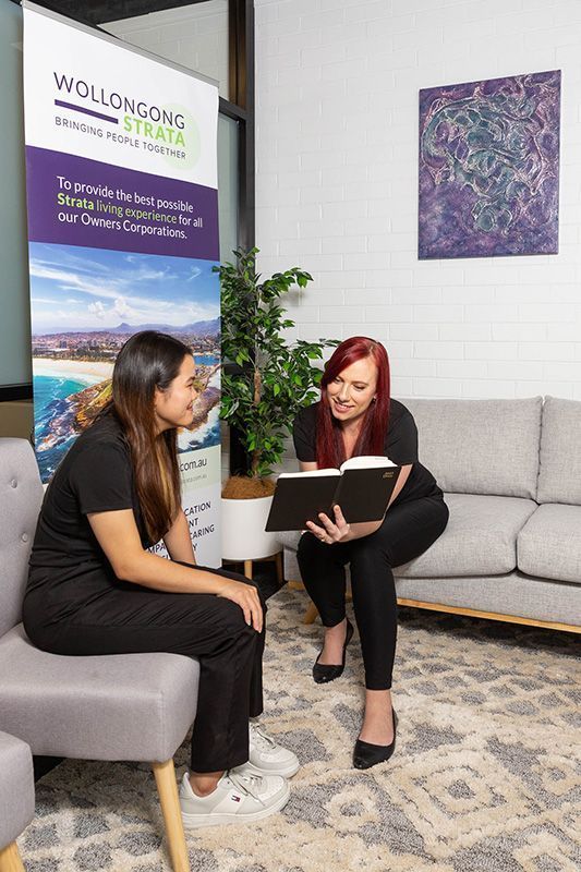 Two women, one with red hair, looking at a book in an office.