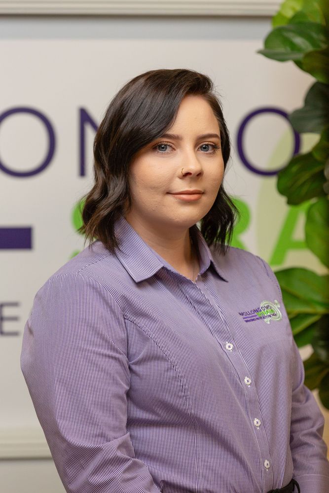 Woman in purple striped shirt smiles at the camera, in front of a logo and indoor plants.