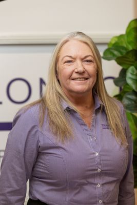 Woman with blonde hair, wearing a purple button-down shirt, smiling, in front of a sign and plants.