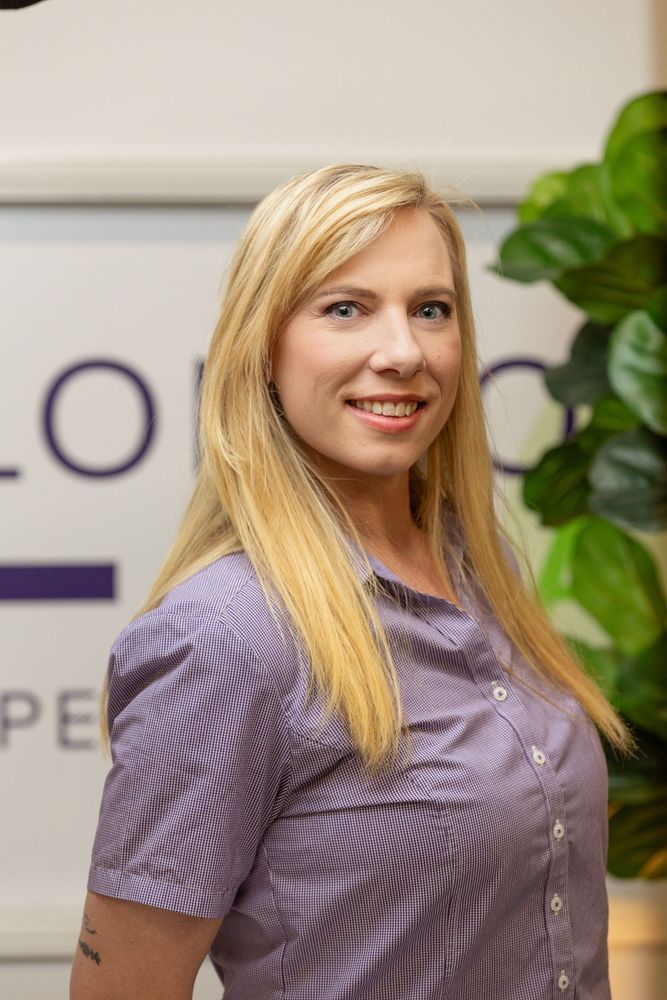 Blonde woman smiling in a purple striped shirt. Background has a logo and plant.