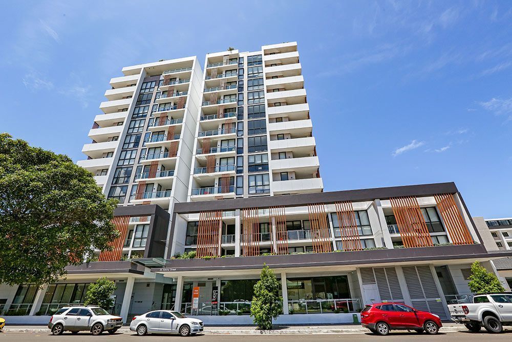 Multi-story modern apartment building with balconies, cars parked in front, and blue sky.