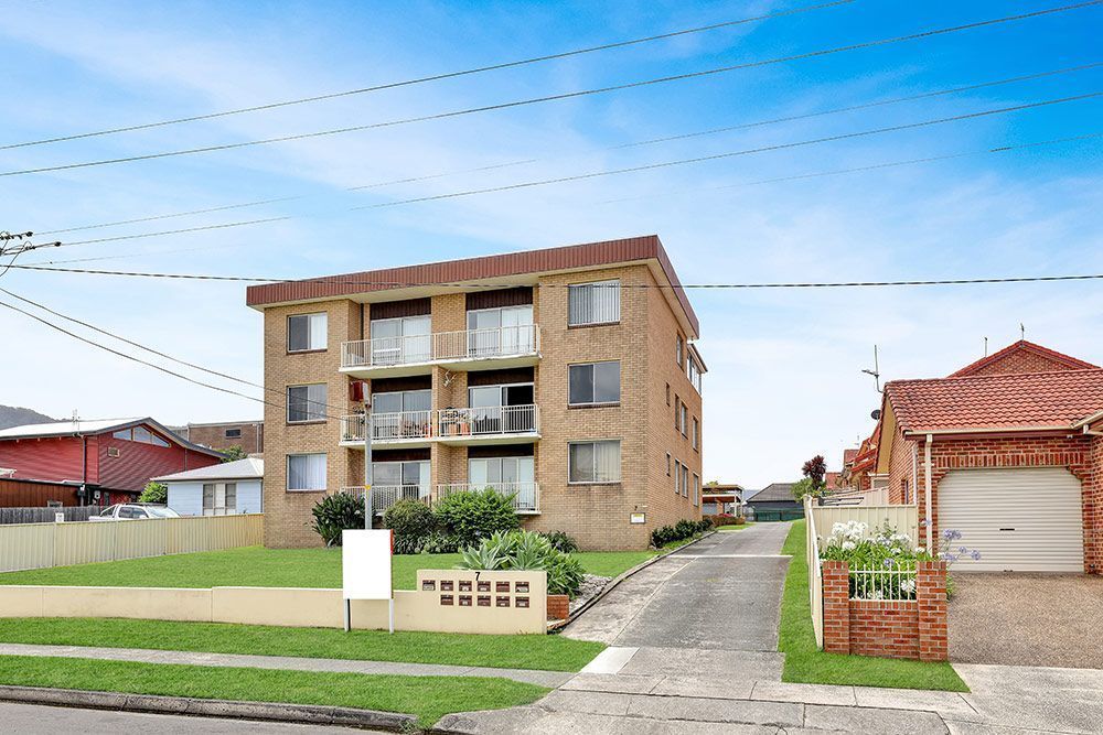 A three-story brick apartment building with balconies, driveway, and grass lawn, under a blue sky.