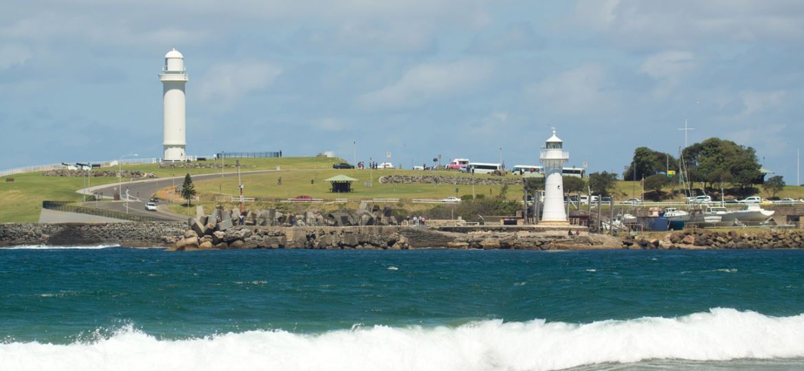 White lighthouses on a grassy peninsula with the ocean in the foreground. Blue sky with clouds.