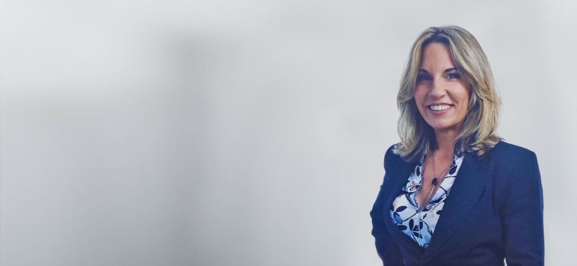 Woman with blonde hair smiles, wearing a dark blazer over a patterned blouse, standing on a white background.