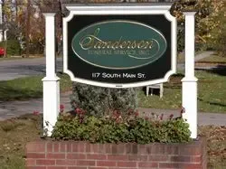 Sign for Sanderson Funeral Service, Inc., at 117 South Main St. with green, gold, and white lettering, set in brick with flowers.