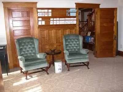 Two green upholstered armchairs face each other, wood-paneled walls, a small table, and a doorway.