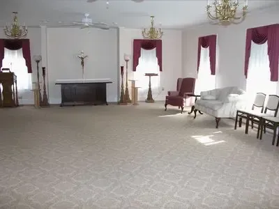 Empty chapel with cross, chandeliers, carpet, windows, and chairs.