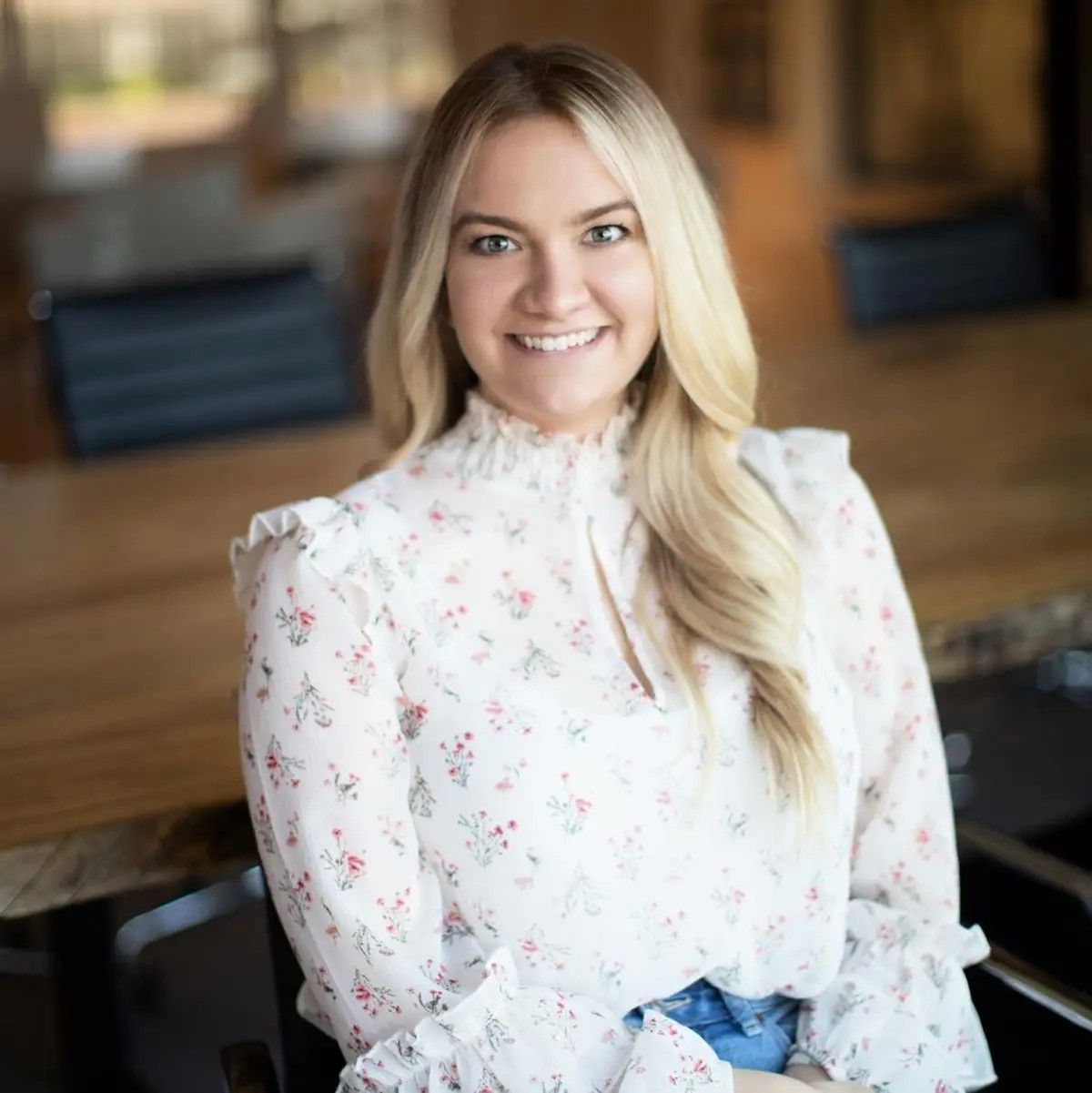 Smiling person with long blonde hair wearing a high-neck floral blouse, seated in a blurred office setting.